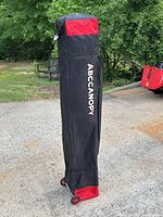 Photo of a black and red wheeled carrying case for a 10x10 ABCCANOPY pop up tent outdoors on a driveway with greenery in the background.