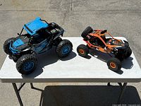 Both radio control cars placed on a white outdoor table photographed in sunlight, showing one blue and black and one orange and black car.