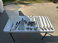 Overview of all items including plastic measuring bucket, whisks, ladles, and forks laid out on a white folding table outdoors.