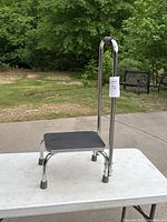 Single step stool with handle, showing side angle view on white table outside with grass and benches in background.