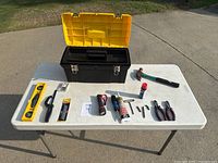Photo of all tools arranged outside the open toolbox on a white folding table, includes hammer, level, saw, flashlight, pliers, screwdrivers, etc.
