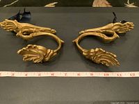 Pair of heavy gilt metal curtain tie backs or hooks, ornate leaf and scroll design, placed side by side with measuring tape showing length.