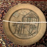 Wide view of charger on patterned rug showing hand-painted naive style house, stick figure, and flower design on center; measuring tape shows about 21 inch diameter.