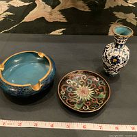 Image showing three cloisonné items: a blue ashtray, a decorative black plate with colorful floral patterns, and a small blue and white vase, all placed on a black surface with a measuring tape.