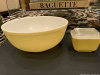 Large yellow Pyrex bowl and smaller yellow square Pyrex dish with clear lid placed side by side on a patterned surface with books in the background.