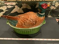 Side angle view of the vintage bird tureen showing the detailed multicolored bird lid on green base, alongside measurement tape for scale.