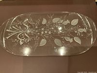 Top view of oval clear glass platter with detailed etched floral and ribbon designs, placed on a dark surface.