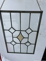 Rectangular stained leaded glass window panel held against a white backdrop showing geometric diamond pattern with amber diamond center and hanging chain.