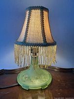 Photo of table lamp turned on with fabric fringe shade, glowing and showing lamp base details
