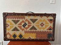 Front view of rectangular suitcase covered in multicolored geometric burlap pattern with leather trim and brass studs.