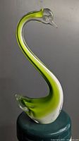 Front view of green and white abstract swan glass sculpture on dark round pedestal, showing smooth curves and glass clarity
