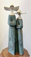 Front view of the two-nuns Lladro porcelain sculpture showing their cream headpieces and greenish-blue robes, standing on a wooden stool.