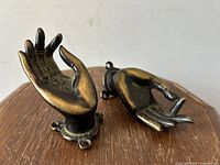 Pair of bronze Buddha hand shaped wall hooks or drawer pulls shown resting on a wooden surface, displaying the detailed front of each hand.