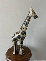 Giraffe statue made of Raku pottery with colorful patchwork design, shown standing on a hexagonal wooden base against a plain wall.