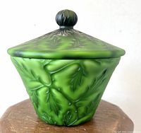 Front view of the emerald green cased satin leaf art glass lidded bowl with embossed leaf design and round finial knob.
