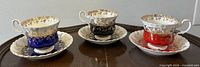 Front view of all three teacup and saucer sets displaying blue, black, and red color designs with gold floral accents on a wooden table.
