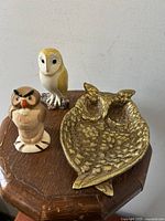 Three owl collectibles on wooden surface: brass owl tray, brown Beswick owl, white and tan Highbank Scotland owl figurine.