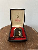 Black Ronson Varaflame Starfire lighter with floral decoration displayed open in original case with red velvet lining on wood surface.