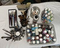 Top view showing multiple paintbrushes stored in circular and rectangular holders, several plastic bottles of acrylic paint, tubes of watercolor paint, and a can of Art Resin