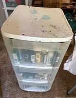A three-drawer plastic storage unit with various paint bottles visible inside, with paint stains on the top surface.