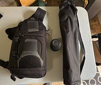 Black nylon shoulder camera bag, small 18-55mm lens and tripod case on white table