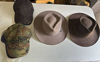 Overview of all five hats - two men's fedoras (light and dark brown) and three baseball caps in various colors (black, camouflage, dark blue)