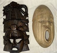 Two wooden masks side by side on a white background, showing Mexican mask with carved pyramid and animal figures and Colombian mask with smooth, rounded features.