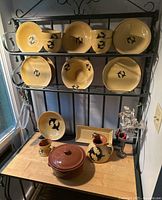 Full view of orca-themed pottery set including plates, bowls, crock pot, pitcher, rectangular dish, and ceramic feathers holder arranged on black metal rack and wood surface