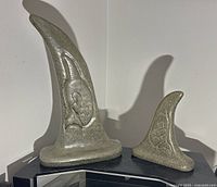 Two polished orca fin sculptures side by side on a black surface, showing overall shapes and intaglio designs