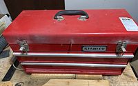 Closed red Stanley metal tool box with black handle and silver latches on wooden surface.