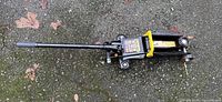 Side angle photo of black trolley jack with yellow safety handle and operational stickers on asphalt surface