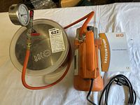 Overall layout of the vacuum pump, vacuum chamber with gauge, and user manual on a white surface, showing the orange pump and hose attached to stainless steel chamber lid.