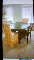 Four chairs around a wooden dining table in a room with window light. Two are black wooden chairs and two are gold brocade upholstered parlor chairs with slipcovers.