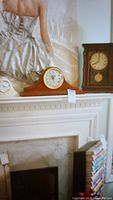 Three clocks placed on a white mantel and shelf, with a painting as background. The mantle clocks include a wooden clock in middle, a ceramic clock in front closeup, and a wooden pendulum clock on the right.