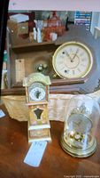 Three clocks on a wooden surface including a Terry Clark antique clock in a basket, a brass Schultz dome clock with glass dome, and a plastic Lorus clock.