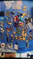 Top view showing an assortment of about fifty earrings laid on a blue surface. Various colors including red, orange, yellow, teal, black and gold-tone. Includes brands Monet, Cato, Joan Rivers, Nina Ricci, Friends collection by Gerald Miller. Mixed clip-on and pierced styles. Photos highlight condition and variety.