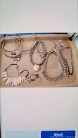 Nine assorted necklaces on a beige tray including silver hoop collars, multi-strand silver necklaces, a stone pendant necklace, and a silver necklace with sparkling stones.