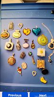 Photo showing many pins and pendants spread on a blue surface. Highlighting various shapes and stone insets including glass hearts and a large filigree pendant.