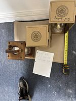 Top-down view of two boxed clear glass oil lamps and one wooden candle lamp on cardboard with tape measure and handwritten note.