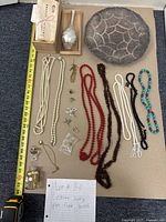Overview of complete lot including costume jewelry, decorative plate, flask, and bottle with handwritten lot note and measuring tape for scale.