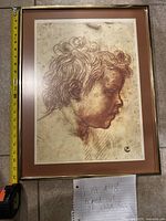 Framed artwork showing a side profile of a child's face with curly hair, brownish sketch, aged paper, brown mat and wooden frame, with measuring tape next to frame for size reference.
