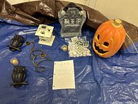 Photo showing all items in the lot arranged on a blue tarp: pumpkin lamp, RIP tombstone, owl candle holders, white lantern, black candle holders, assorted LED candles.