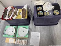Photo showing all plates and accessories in the lot arranged on plastic storage bins, including round multicolor plates, brown elongated trays, two white fish-shaped plates and ceramic stands.