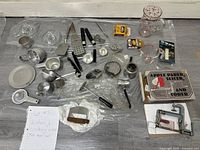 Overhead view of assorted kitchen and cooking ware laid out on the floor showing utensils, graters, peelers, measuring cups, strainers, and boxed apple parer tool.