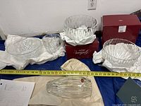 Wide view of large crystal bowls and vases arranged with two red Brilliance Collection boxes, measuring tape showing size scale, some items wrapped in cloth or packaging indicating new or gently used condition.