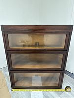 Front view of the wooden display shelf with three glass-front compartments, measuring tape placed for scale