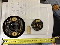 Top-down view of the black lid with gold floral design next to a measuring tape and handwritten note, showing size and condition with visible crack.