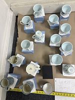 Top down view of 16 white ceramic mugs, some with blue floral designs and some with blue polka dots, each mug placed on original blue packaging boxes with tags.