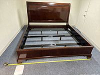 Full view of assembled wooden bed frame without mattress inside a room, showing headboard, side rails, and footboard. Yellow tape measure is in the foreground to indicate size.