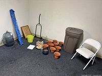Photo showing the entire grouping of items including dolly, folding chair, terracotta pots, glass bottles, yellow bucket, framed item, trash can, and a blue-striped folded umbrella.